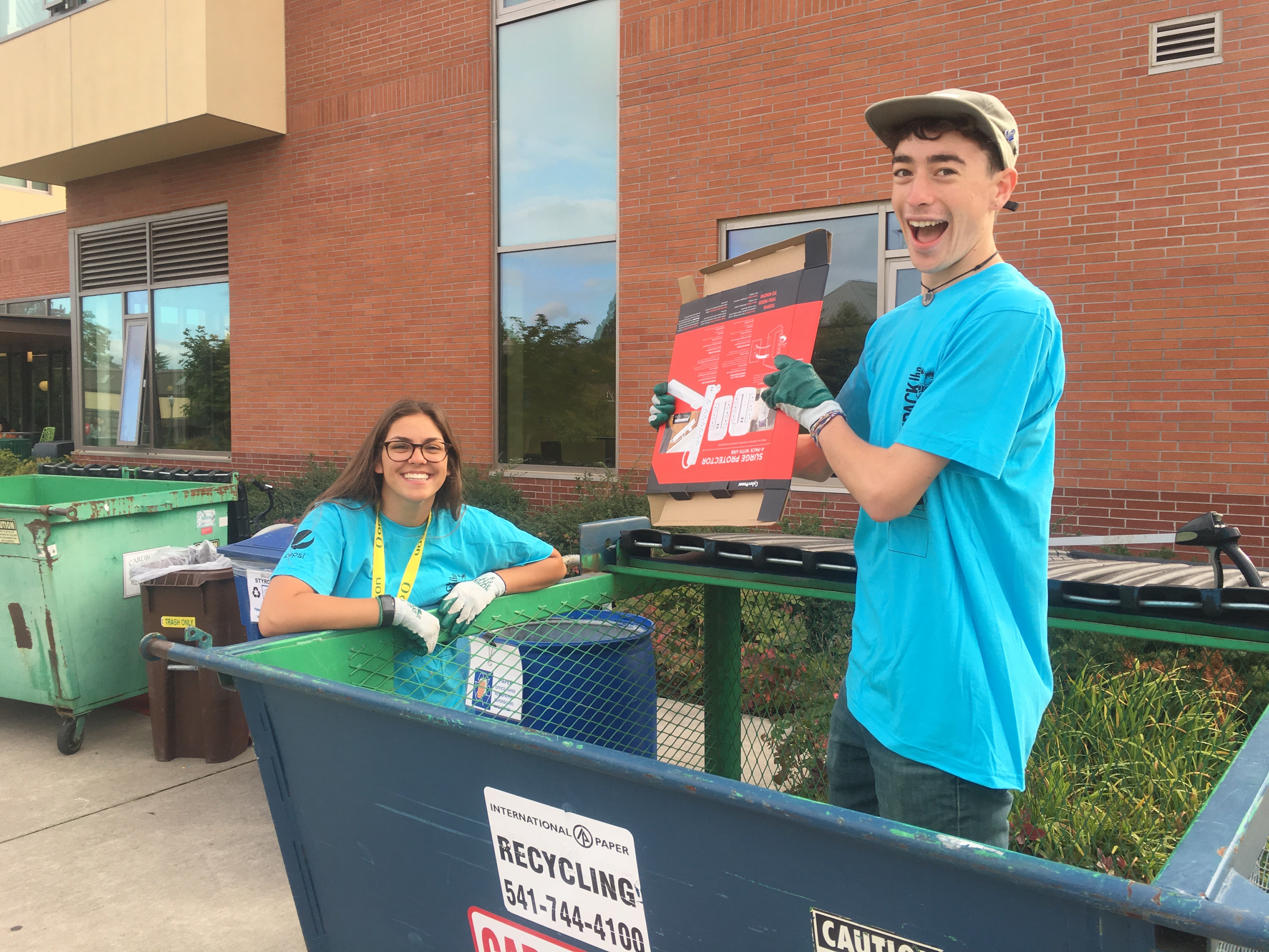 A female and male student stand in a dumpster and show broken down cardboard as part of campus recycling efforts.as part of 