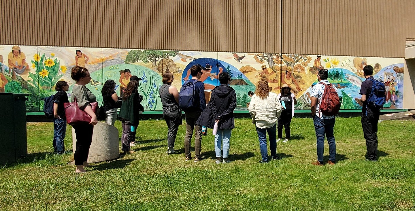 UO Sustainability Fellows learn from community partners in front of a Kalapuya mural.