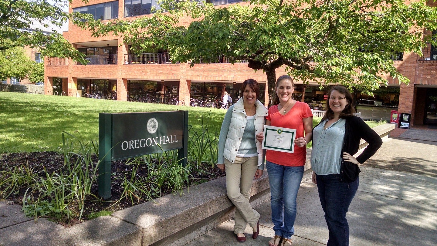 Three UO staff members from the Business Affairs Office stand in front of the Oregon Hall sign holding their Green Office certificate.