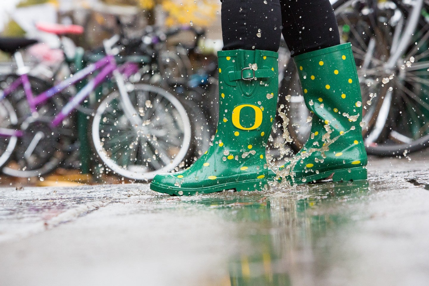 Rainboots with the UO "O" in a rainy puddle with bicycles in the background.