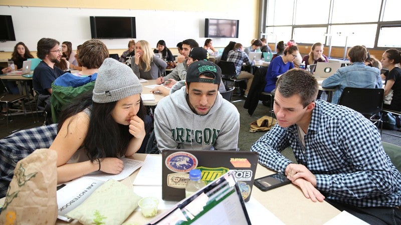 Three students work on a group project in a classroom with other students working as well.