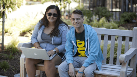 Two students sit and smile on a bench on campus.